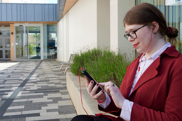 A person sitting outside the Anne Rowling Clinic building dressed in a maroon jacket with headphones on and scrolling on a mobile phone