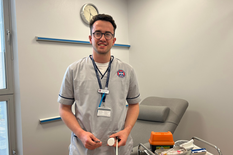 A picture of Calum Scoular taken inside one of the Anne Rowling Clinic's clinical rooms. Calum is wearing a University of Edinburgh student nursing uniform and is holding a tendon hammer. Behind Calum is an examination couch