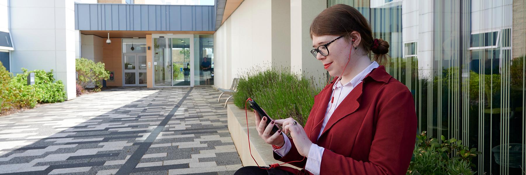 A person sitting outside the Anne Rowling Clinic building dressed in a maroon jacket with headphones on and scrolling on a mobile phone
