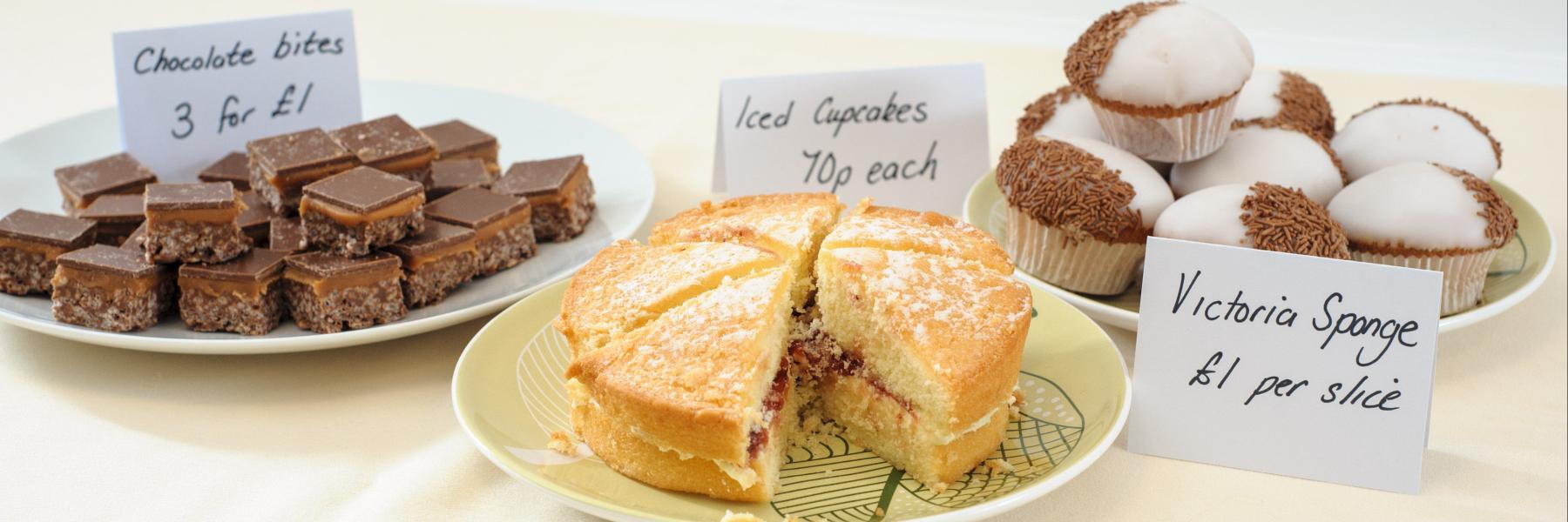 Image of a bake sale with chocolate bites (right), Victoria sponge cake (centre) and chocolate cupcakes (left)
