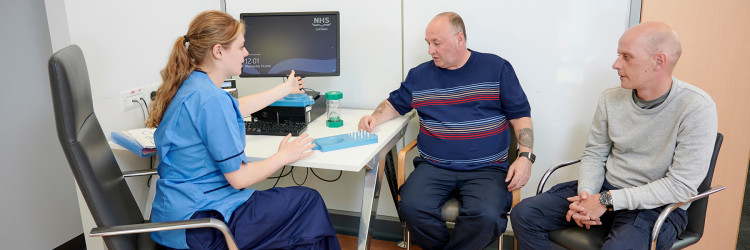 An NHS research practitioner is in a clinic room with two men. One is performing a cognitive test, moving pegs on a plastic board, while the other looks on.