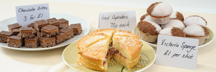 Image of a bake sale with chocolate bites (right), Victoria sponge cake (centre) and chocolate cupcakes (left)
