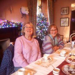 A picture of fundraiser Linda McTurk at a table laden with lots of cakes and baking, the setting is Christmas and there are sparkling lights in the background. Linda and her friend are smiling at the camera while enjoying a cup of tea.