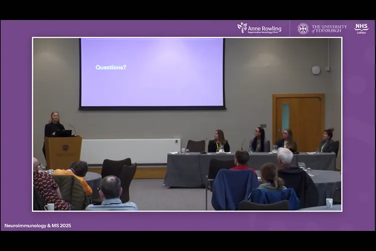 screenshot from the event recording showing a speaker at a lectern, 4 panel members at a table, and several people in the audience.