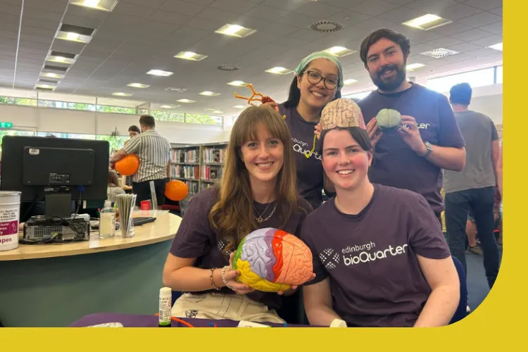 A picture of Jade Lucas with three researchers at a science communication event. One researcher is wearing a hat styled like a brain and another researcher is holding a coloured model of a brain. On the table in front of the researchers are craft materials.